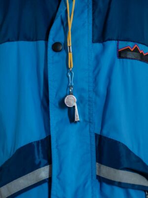A close-up of a blue jacket featuring a whistle and lanyard, showcasing outdoor apparel.