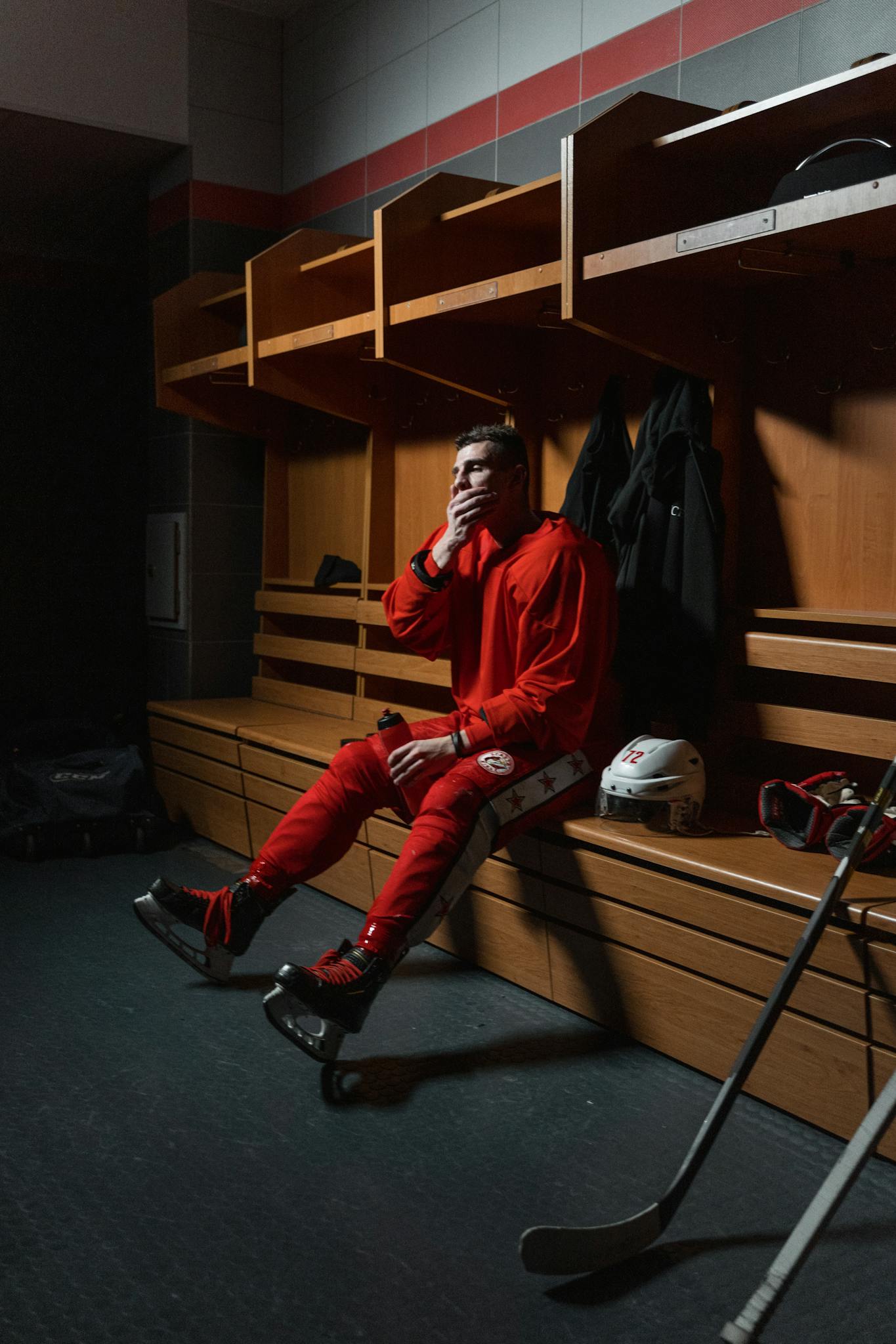 A hockey player in red uniform sits thoughtfully in a dimly lit locker room.